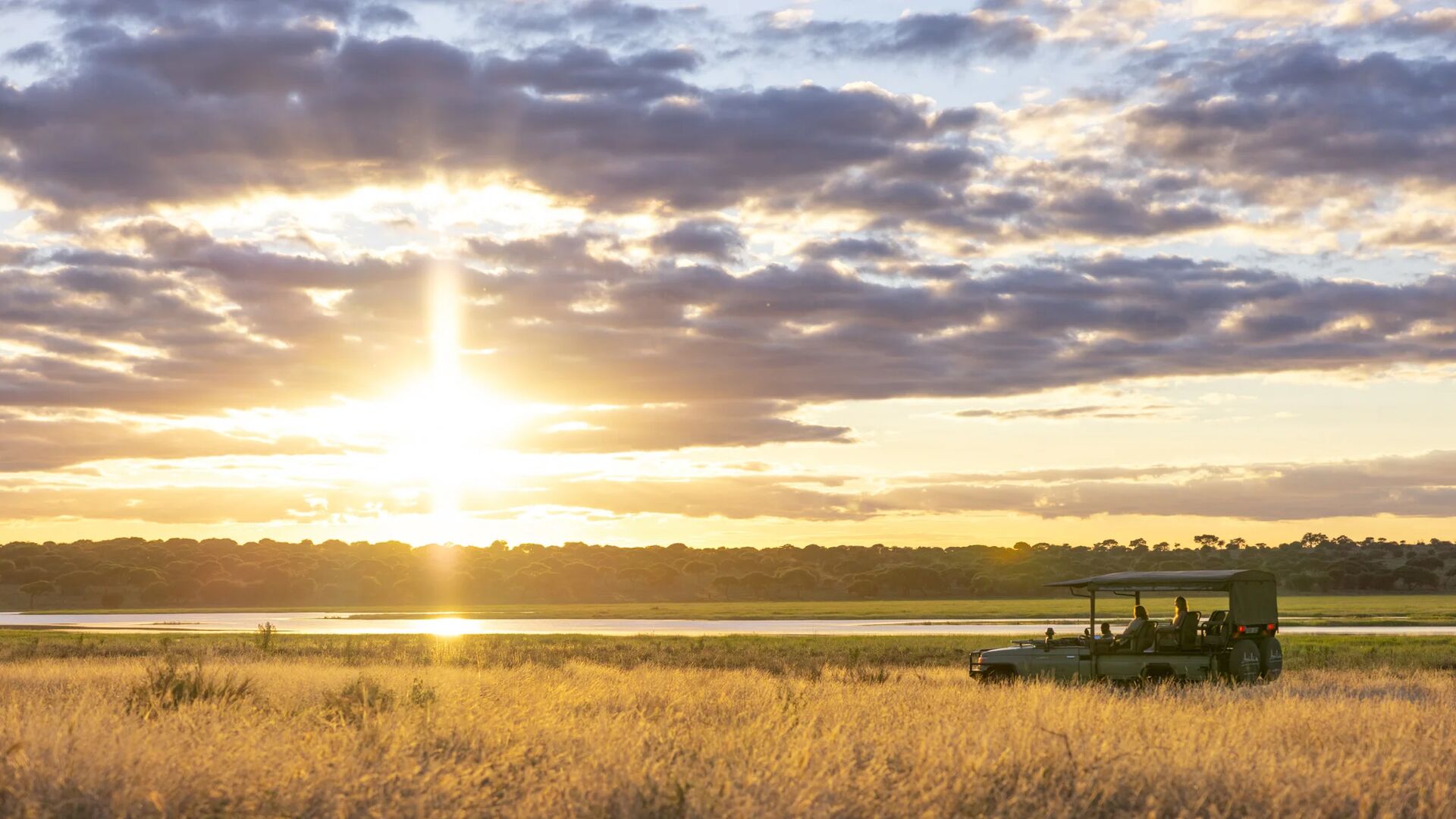 Oliver's Camp, Tarangire, an open safari vehicle carrying guests pauses in tall golden grass at sunset, overlooking a calm waterway as dramatic clouds and warm sky light