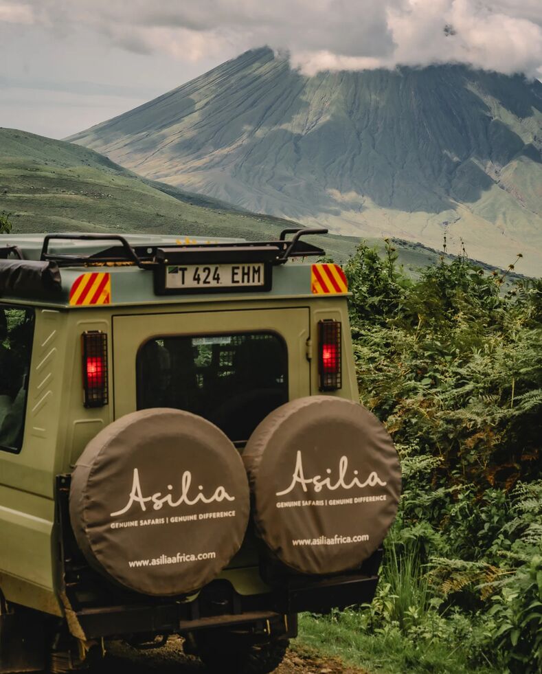 An Asilia safari vehicle parked on a green track with spare wheels visible, facing the steep volcanic slopes of Ol Doinyo Lengai rising under low clouds in the Ngorongoro Highlands.