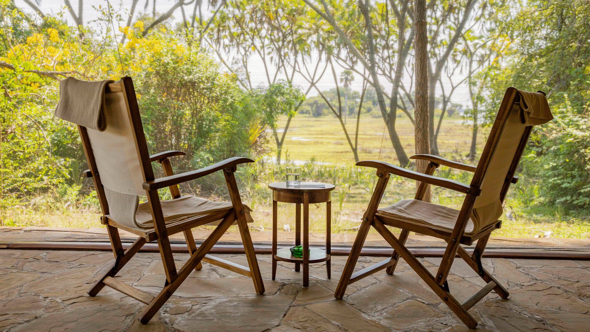 Two wooden chairs and a small table on a deck facing a green riverside view through trees at Roho ya Selous Camp
