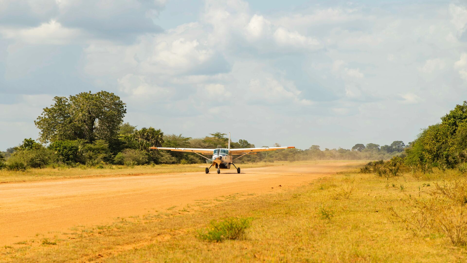 A small propeller plane taxiing on a dusty airstrip surrounded by grassy savannah