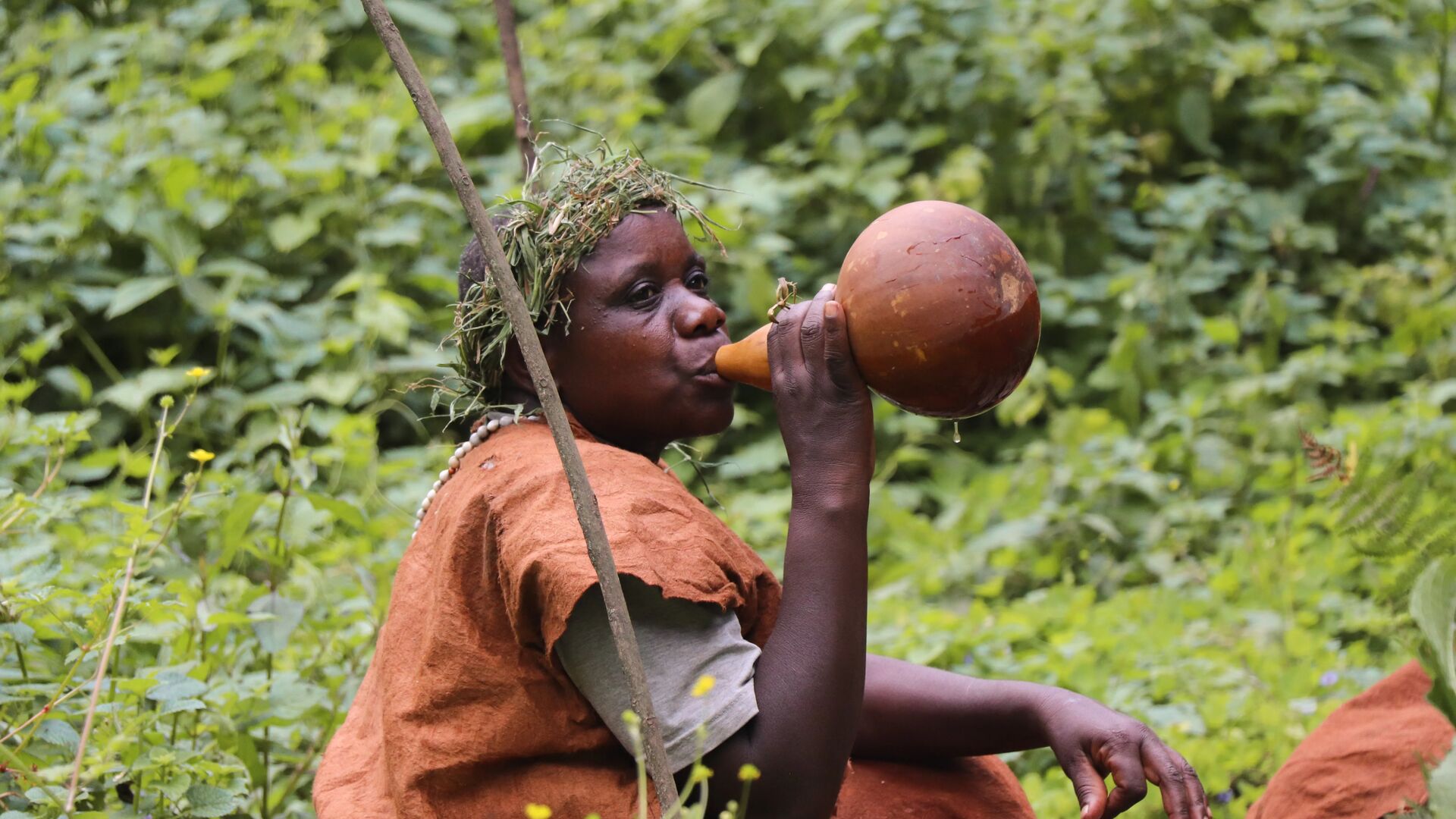 A Batwa woman wearing a grass headpiece and rust-coloured cloth drinks from a traditional gourd amid dense green vegetation in Bwindi Impenetrable Forest, Uganda.
