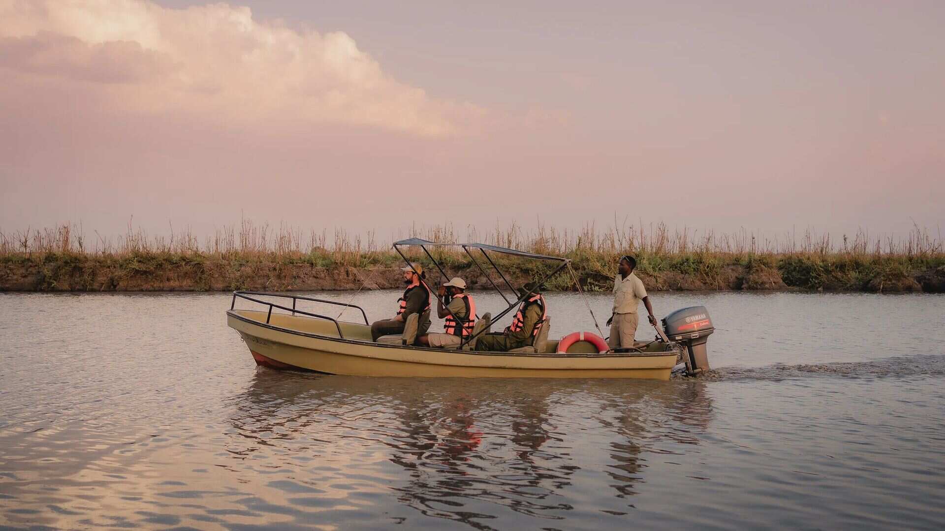 Guests wearing life jackets and a guide cruise along a calm river in a small motorboat under a soft pink sky at dusk, Ruaha National Park, Tanzania.