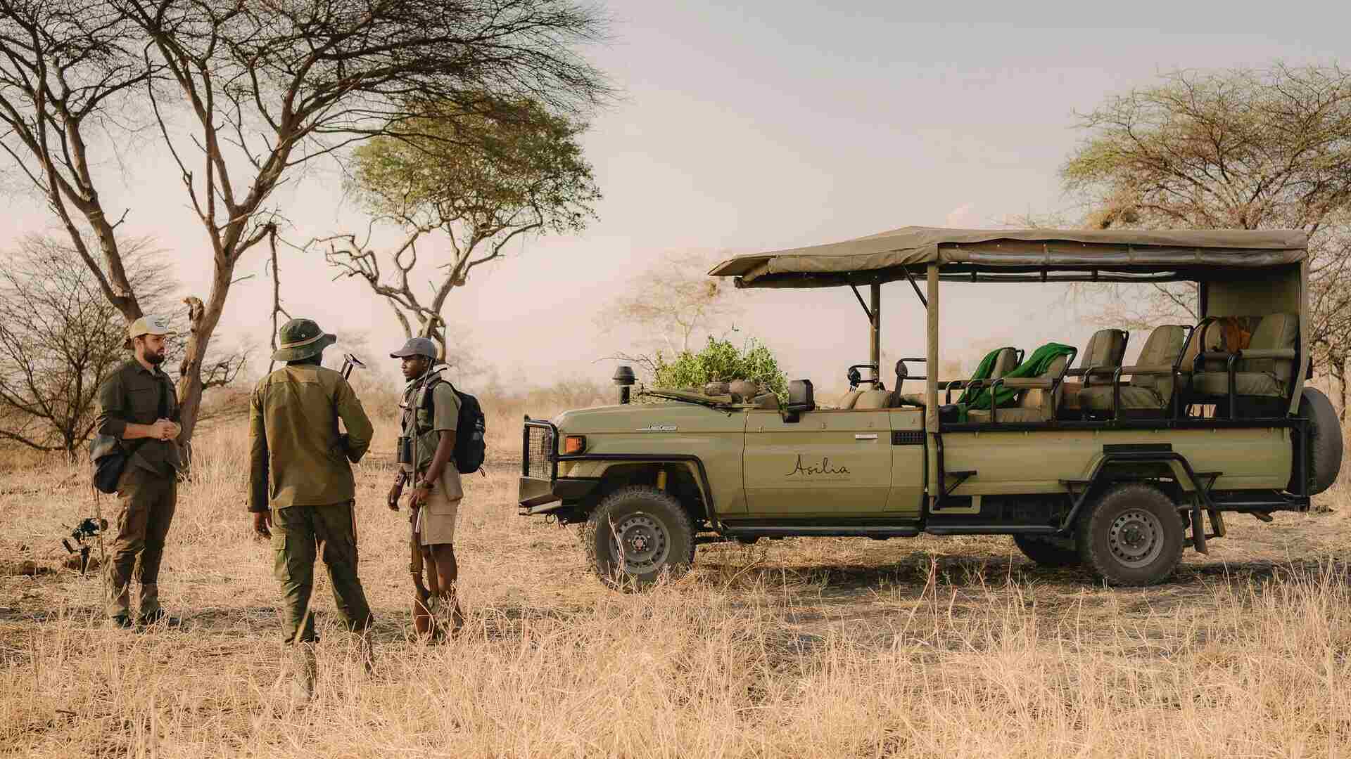 A guest and two Asilia Africa guides stand beside an open-sided safari Land Cruiser in dry golden bush dotted with acacia trees, Ruaha National Park, Tanzania.
