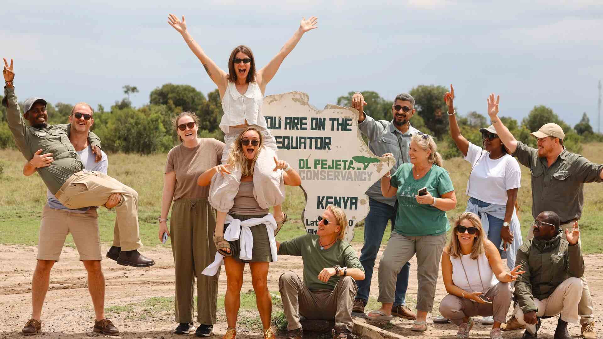 A lively group of guests and Asilia Africa guides pose laughing and cheering in front of the "You Are On The Equator — Ol Pejeta Conservancy, Kenya" sign, Kenya.