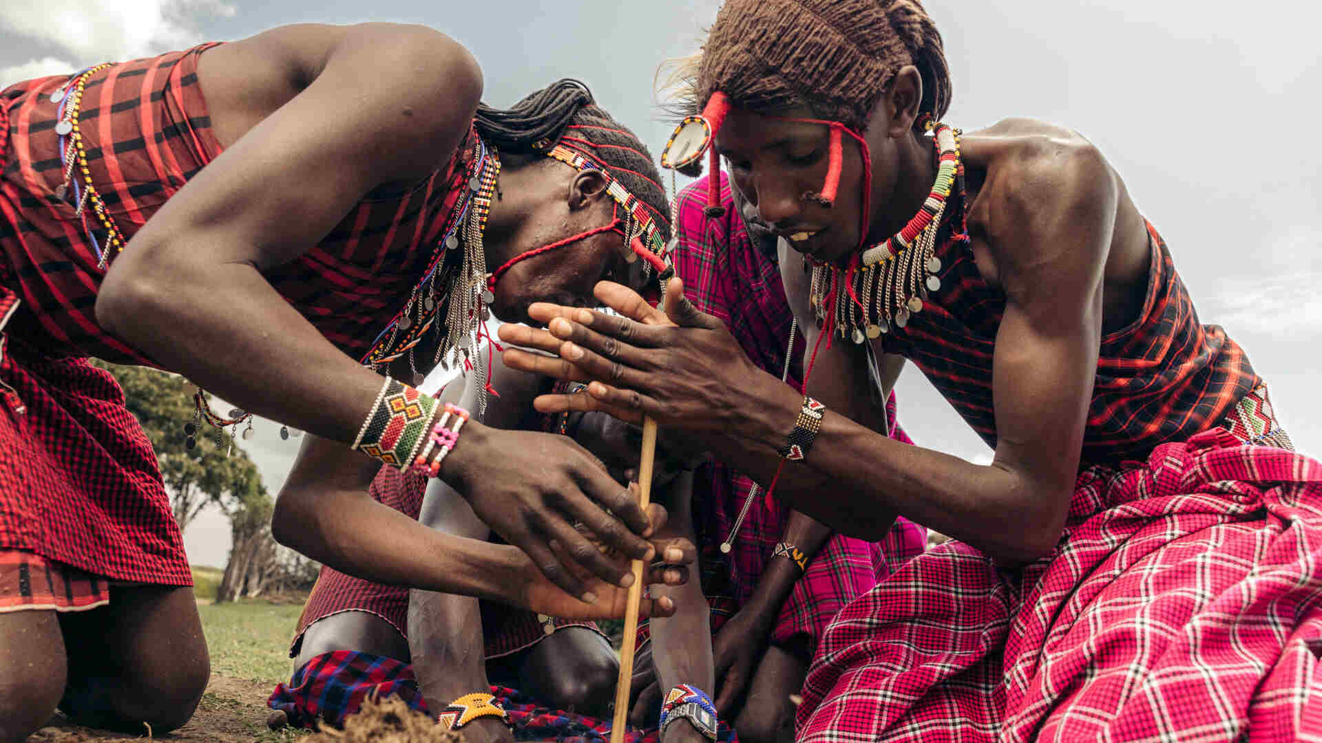Maasai men wearing traditional red and pink shuka wraps and beaded jewellery crouch together to start a fire using a wooden friction stick, photographed near Emboo Camp in Kenya.