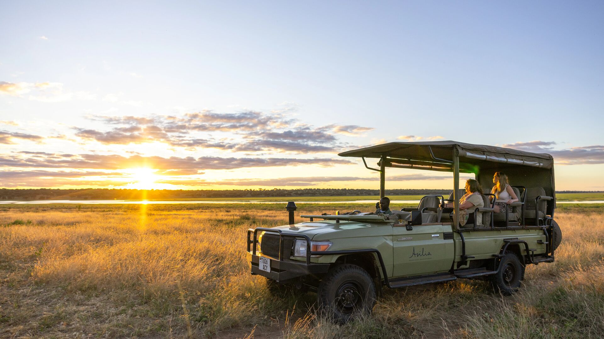 Guests and a guide in an open-sided Asilia Africa Land Cruiser watch the sun set over golden grassland and a distant lake in the Serengeti, near Oliver's Camp, Tanzania.