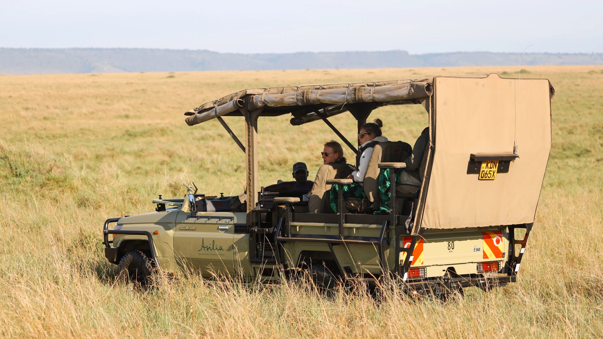 Guests seated in an open-sided Asilia Africa Land Cruiser safari vehicle scan the open Masai Mara plains, with golden grassland and the Oloololo Escarpment visible on the horizon near Rekero Camp, Kenya.