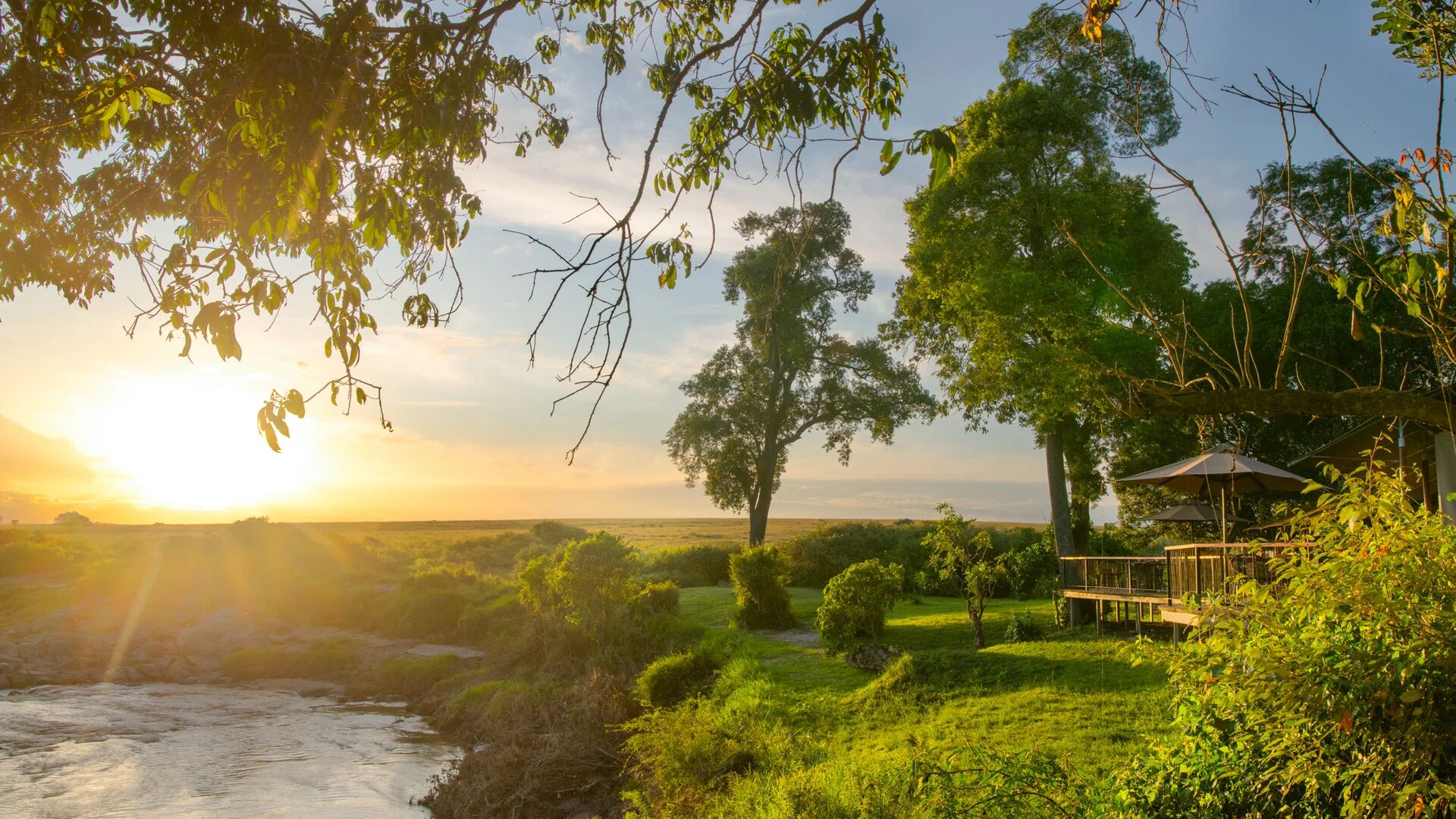 Sunset view from Rekero Camp in the Maasai Mara, with golden light streaming through riverine trees over the Talek River, lush green gardens, and a raised wooden deck with parasols overlooking open savannah plains.