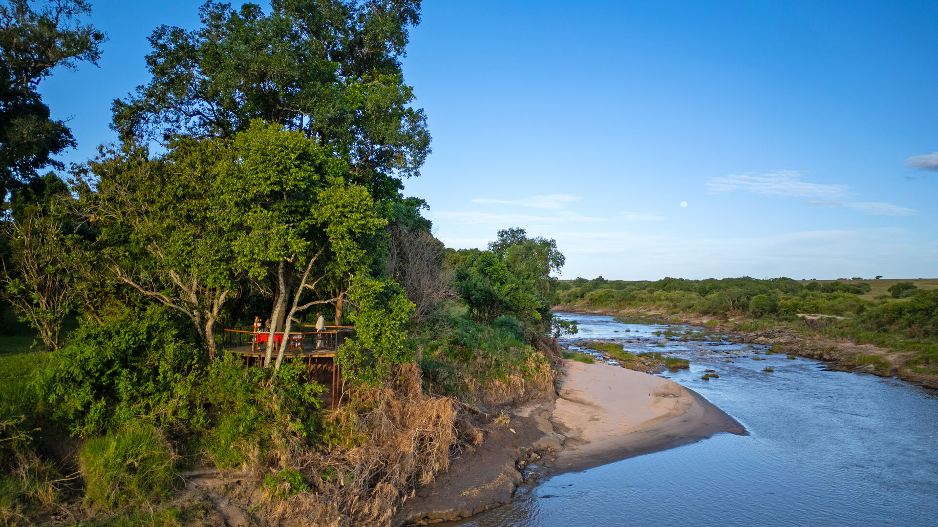 Aerial view of Rekero Camp's raised wooden deck nestled among lush riverine trees on the banks of the Talek River, with sandy banks, shallow rapids, and green bush stretching into the distance.