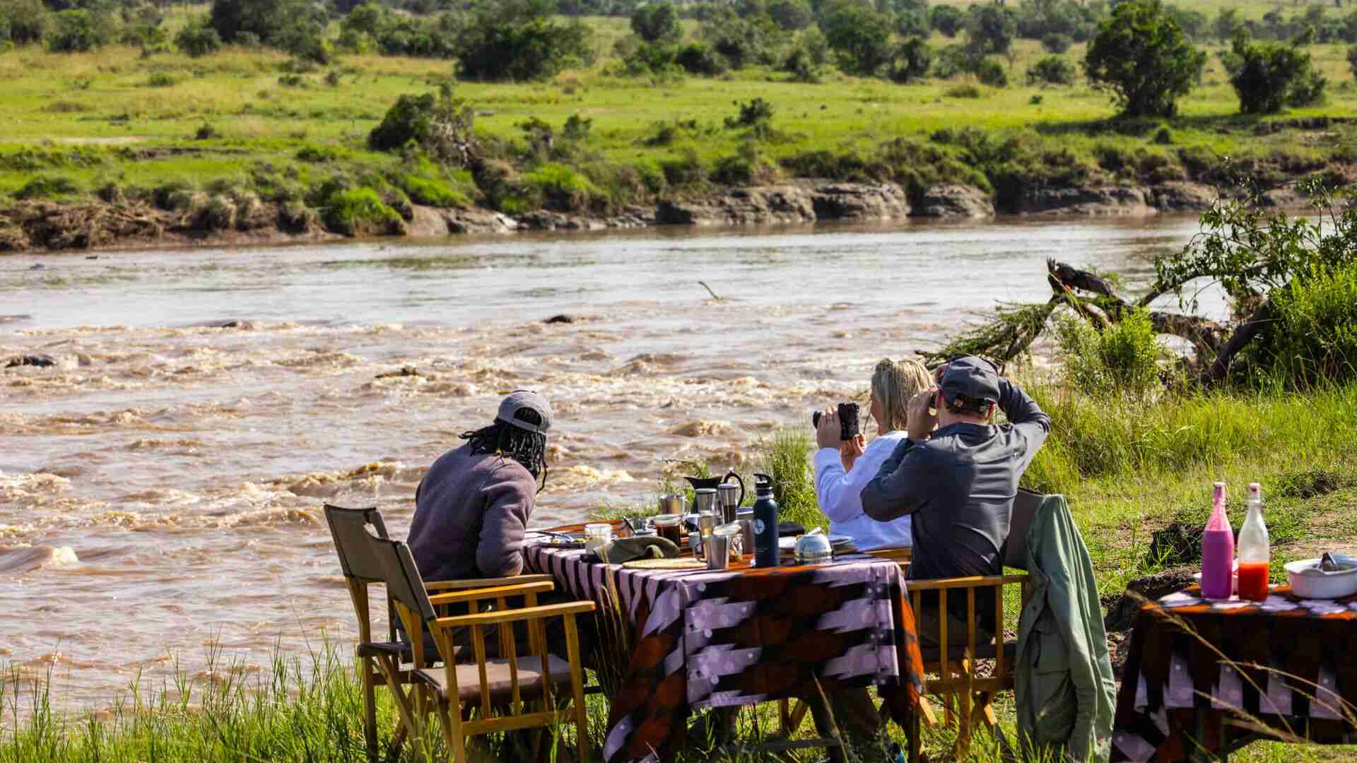 Three guests and a guide sit at a cloth-covered table set up on the grassy riverbank, watching the rushing brown waters of the Mara River through binoculars and a camera, during a bush lunch from Olakira Camp, Serengeti, Tanzania.
