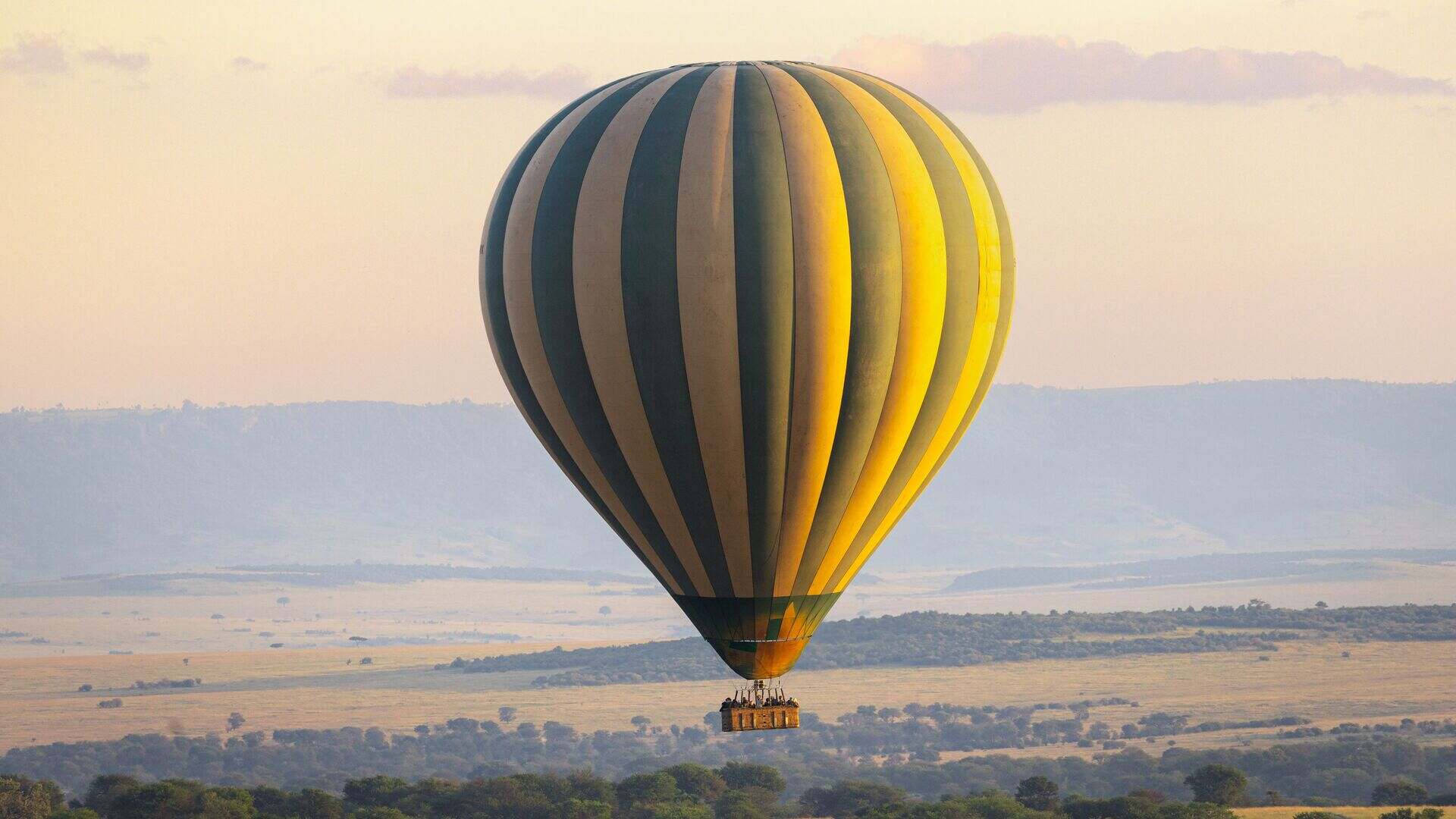 A green-and-yellow striped hot air balloon floats above the vast Serengeti plains at dawn, with hazy savannah and distant hills stretching to the horizon, near Sayari Camp, Tanzania.