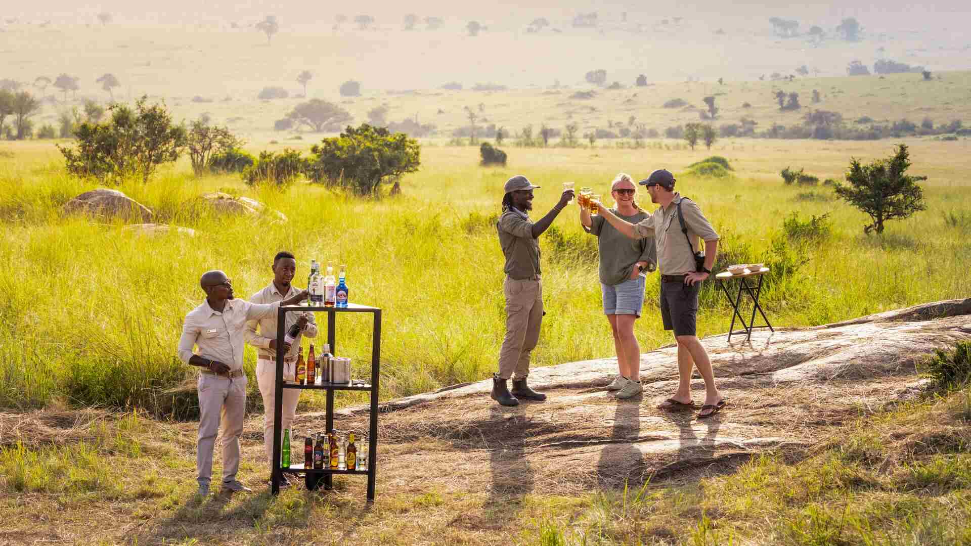 Guests and Asilia Africa staff raise glasses in a toast on a granite kopje with a mobile drinks trolley, overlooking the vast green Serengeti plains at golden hour, near Olakira Camp, Tanzania.